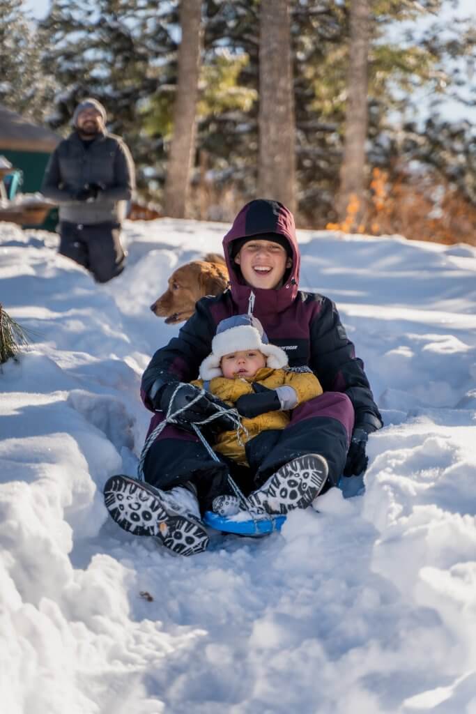 a teenage boy and a toddler in snow gear riding in a snow sled down a  snowy hill with a dog in the background