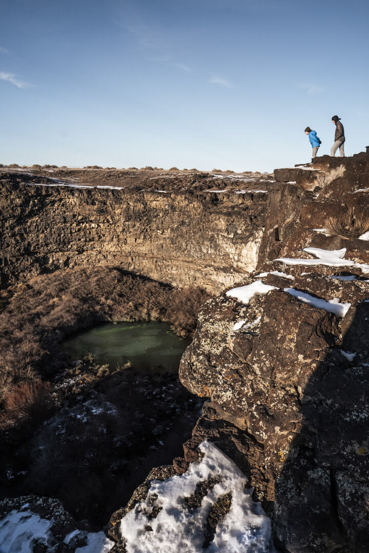 a mom and her son stand on the edge of rocky overlook, looking down at a blue pool of water on the canyon floor