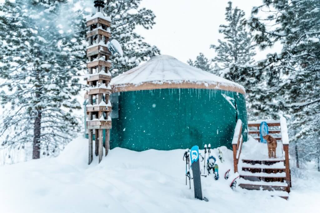 Exterior photo of green yurt with brown roof covered in snow and icicles with snowboards and ski gear standing outside.