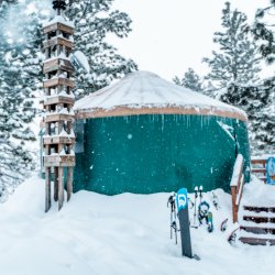 Exterior photo of green yurt with brown roof covered in snow and icicles with snowboards and ski gear standing outside.