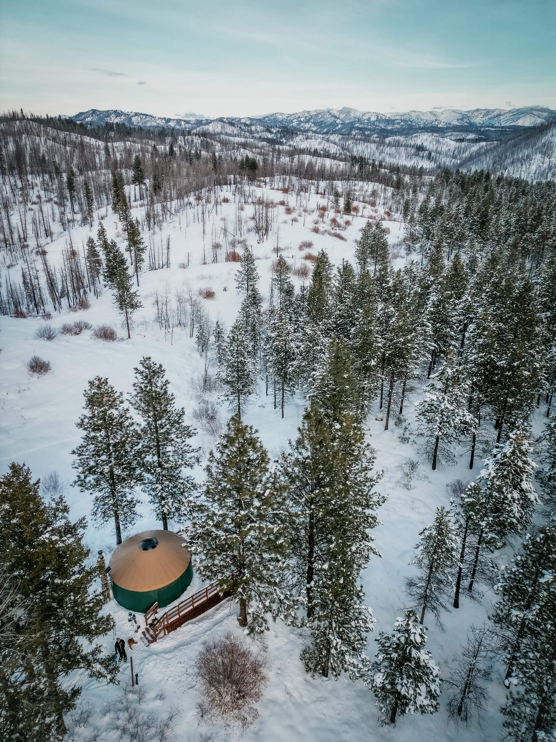 Aerial view of green yurt with brown top surrounded by snow and snowy trees with snow-covered mountains in the distance.