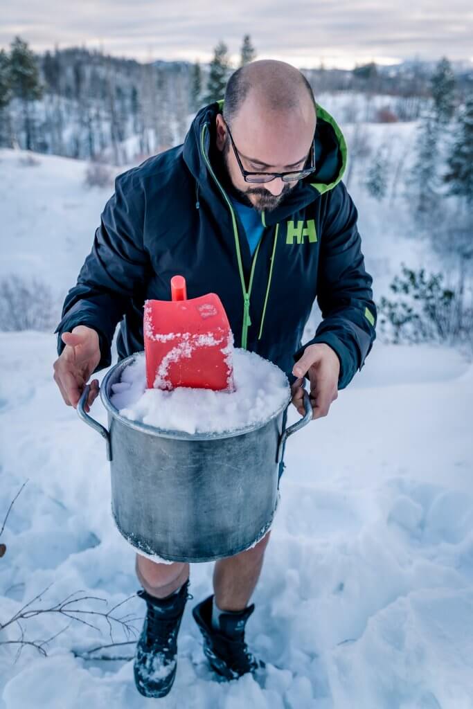  a man in a winter jacket carrying a large silver pail filled with snow and red scoop on top of the pile of snow in the bucket