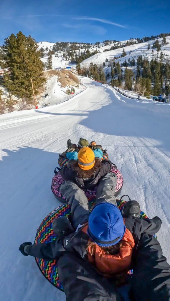 four people on snow tubes in a line holding onto each other's feet going down a snowy snow tube lane at bogus basin