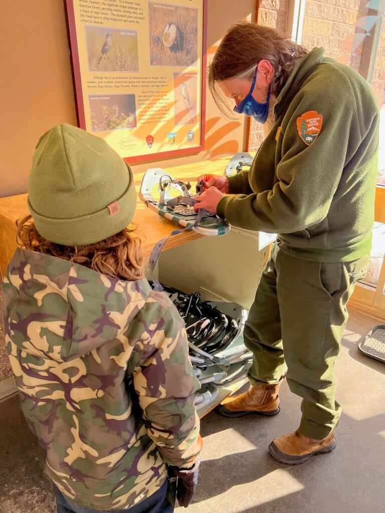 a young boy in a camo snow jacket watching a park ranger prepare snowshoes inside the craters of the moon visitor center