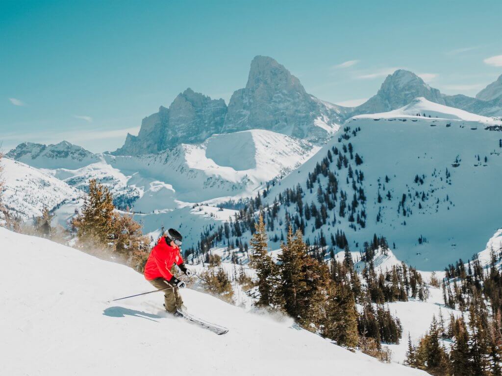 a skier in a bright red coat on snowy slope with rugged Teton Mountains in the background