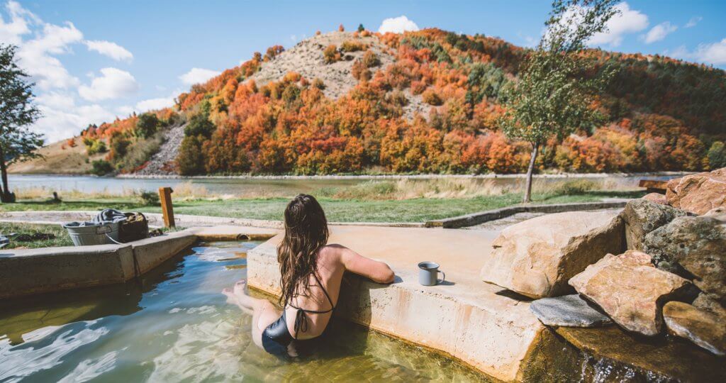 A woman wearing a black one piece bathing suit, sitting in a hot pool at Maple Grove Hot Springs, staring at a hill covered in red, orange, yellow and green foliage.