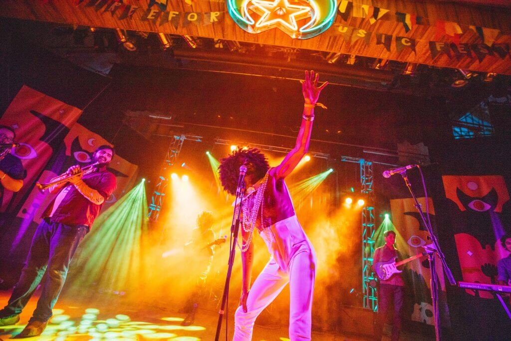 a black singer where high waisted white pants stands at a microphone on an elevated stage. Neon lights are shining down on him and his band members.