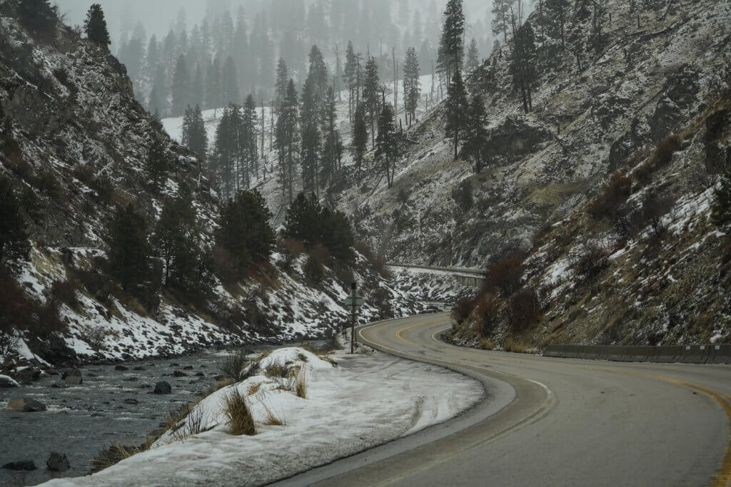 winding road with snowlined river on one side and mountain and snowcovered tress on the other side