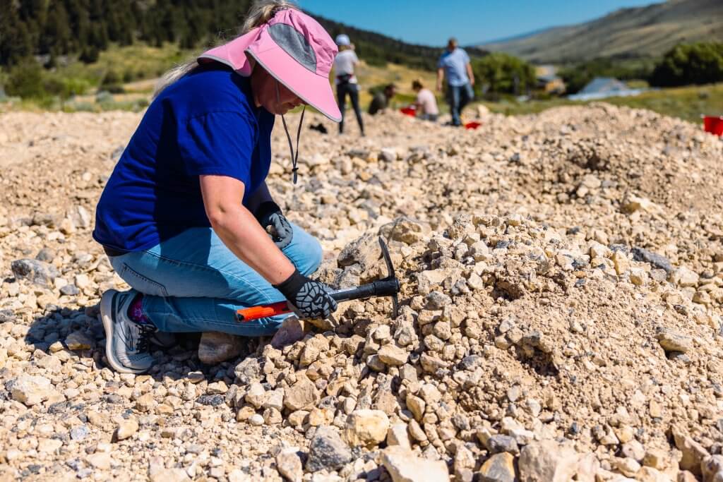 a woman in a t-shirt, jeans and sun hit sits atop a rock pile with rock hammer looking for opals at Spencer Opal Mines