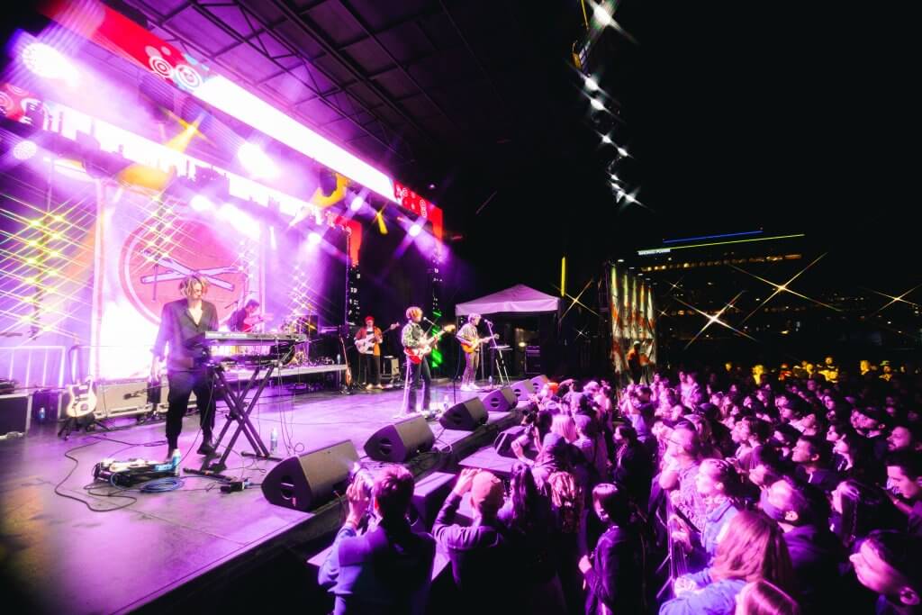 Main Stage at Treefort Music Fest light up by neon purple lights. A guitarist and keyboard player are on stage with a large crowd of people gathering at the bottom of the elevated stage.