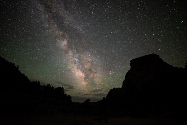 a dark night sky with the Milky Way in the middle of the photo and the dark outline of rock formations along the side