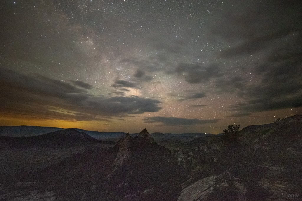 wide night sky shot filled with lots of stars and a few light clouds with shadowed rock formations below