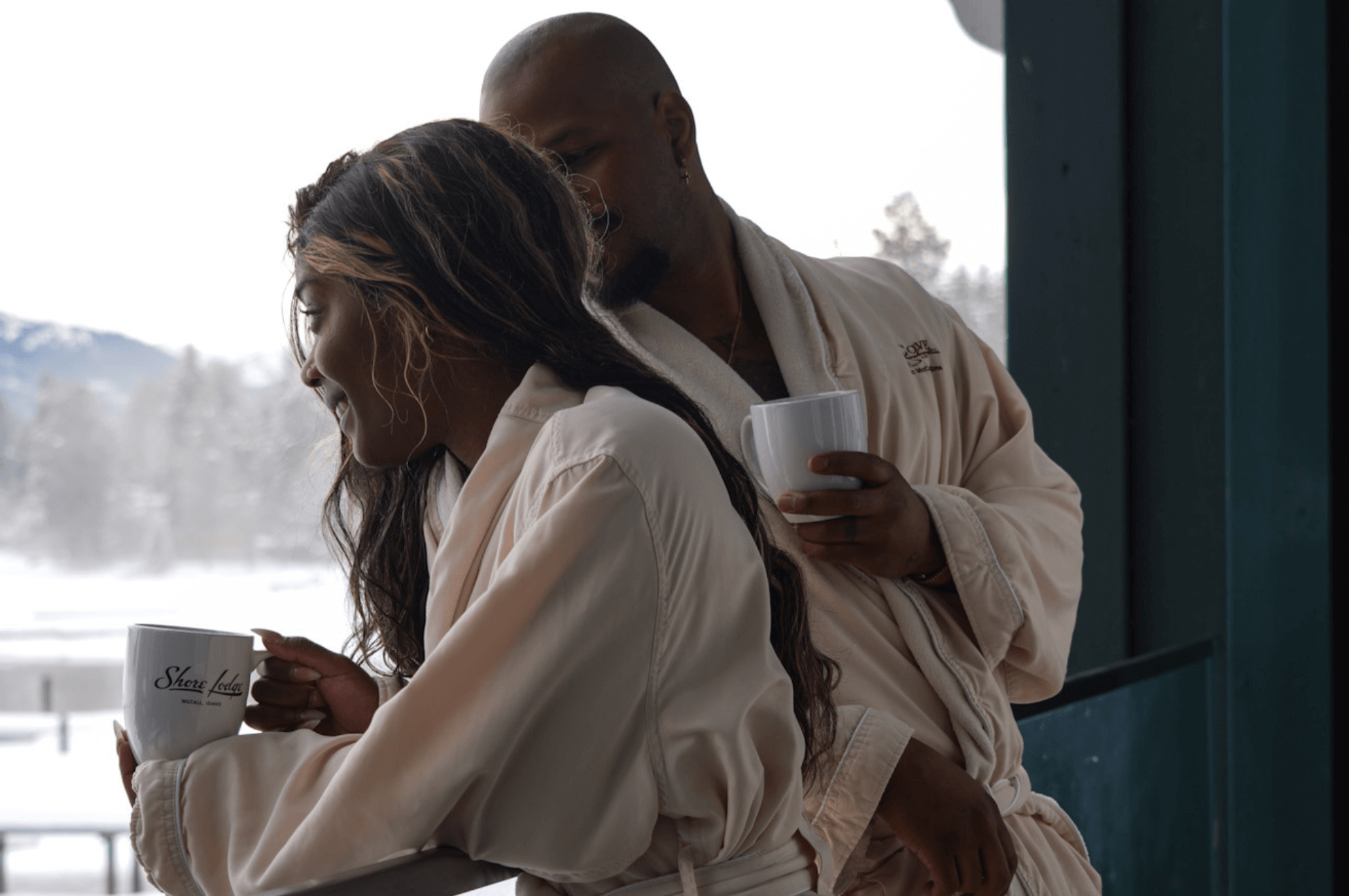 two black adults in white bathrooms holding cups of coffee on a hotel room balcony looking at frozen Lake Payette