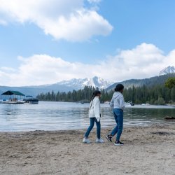 Two girls walk along the sandy shoreline of Redfish Lake with the Sawtooth Mountains and forest in the background.