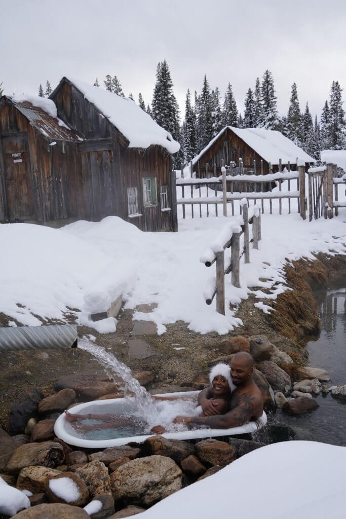 a black man and woman sit outside in a rustic tub where a pipe is pouring water onto them with surrounded by snow and a few small, rustic cabins at Burgdorf hot springs