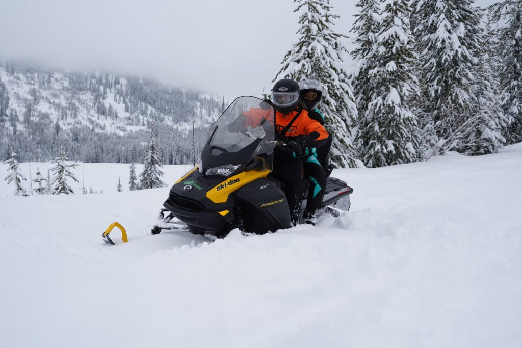 two people on a snowmobile in snow gear sitting in deep snow powder and surrounded by snow-covered pine trees