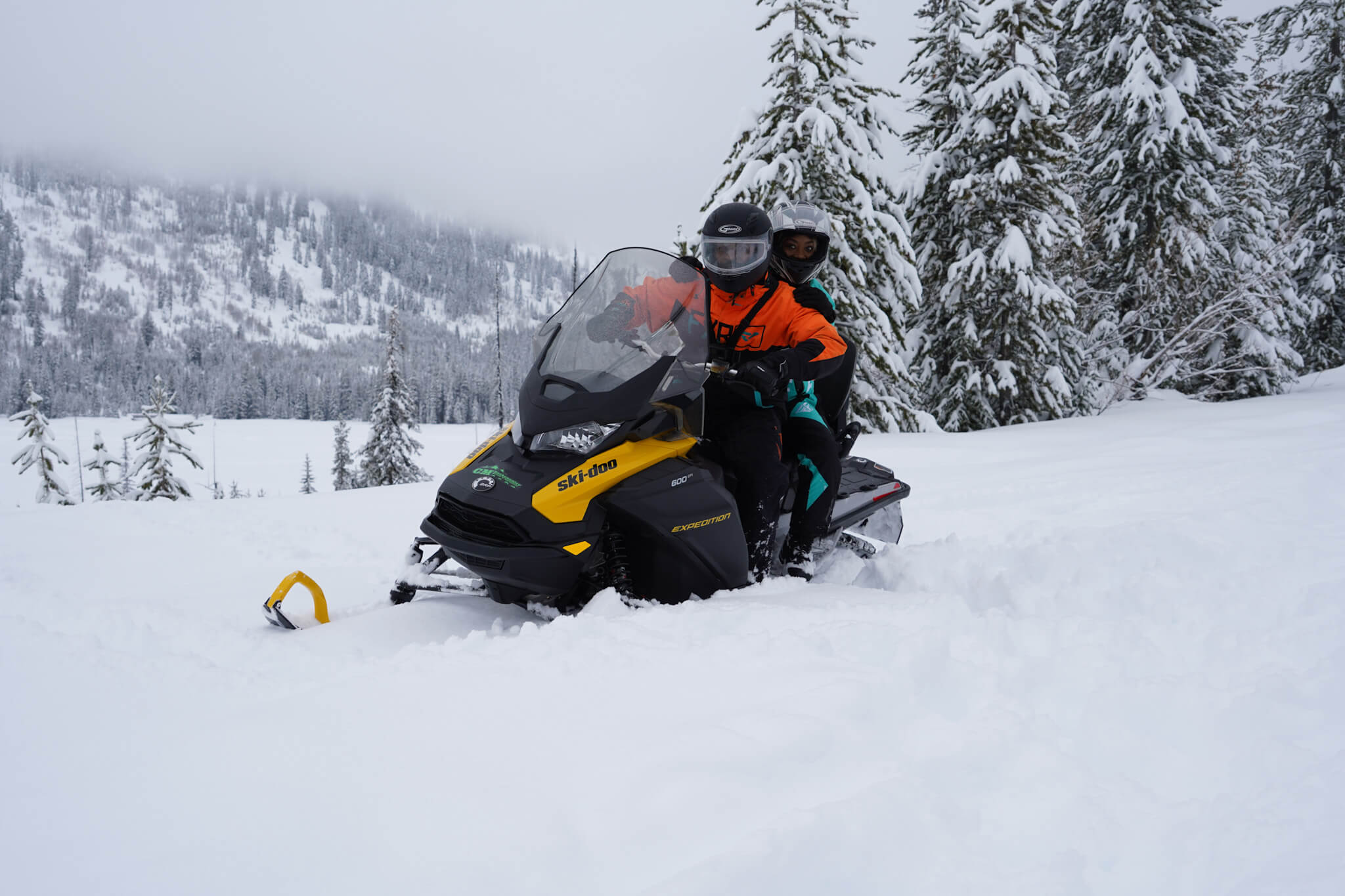two people on a snowmobile in snow gear sitting in deep snow powder and surrounded by snow-covered pine trees