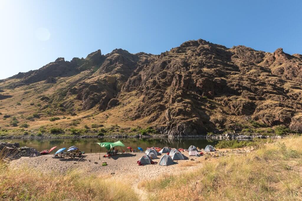 Multiple tents are setup on a sandy beach along the Snake River in Hells Canyon. Rugged hills and sagebrush sit behind the tents.