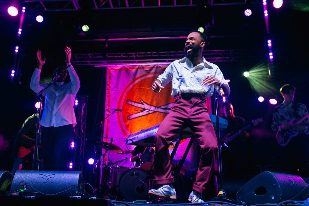 Musician Joshy Soul, dressed in burgundy pants, a light colored shirt, and light colored suspenders, dances on stage in front of a Treefort sign.