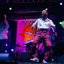 Musician Joshy Soul, dressed in burgundy pants, a light colored shirt, and light colored suspenders, dances on stage in front of a Treefort sign.