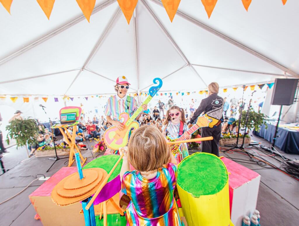 Two children and one adult are dressed in colorful attire performing as a band on cardboard instruments at Kidfort.