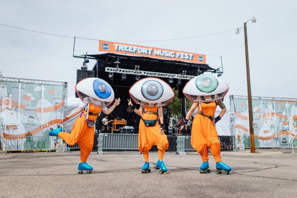 Thee people on rollerskates wearing bright orange rompers and a single giant eyeball on each of their heads. All three rollerskaters are standing in front of an orange Treefort sign.