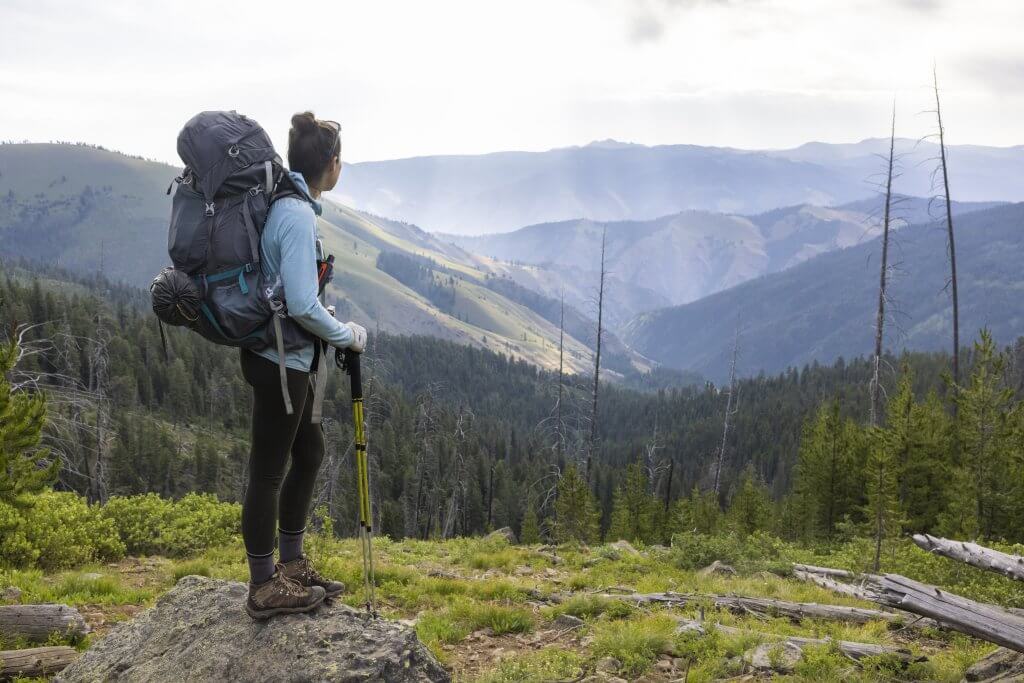 A female hiker stands on a rock wearing a full backpacking pack of gear looking into the distance at the Seven Devils Mountain Range.