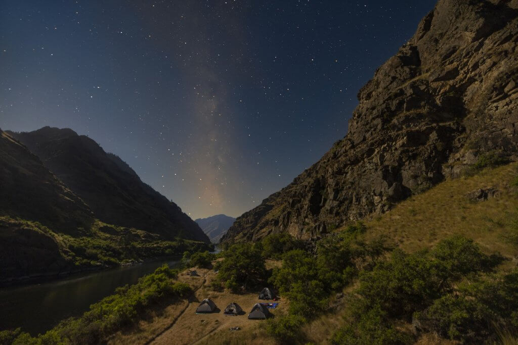 Several camping tents set up on a sandy beach along the Snake River in Hells Canyon under a starry night sky.