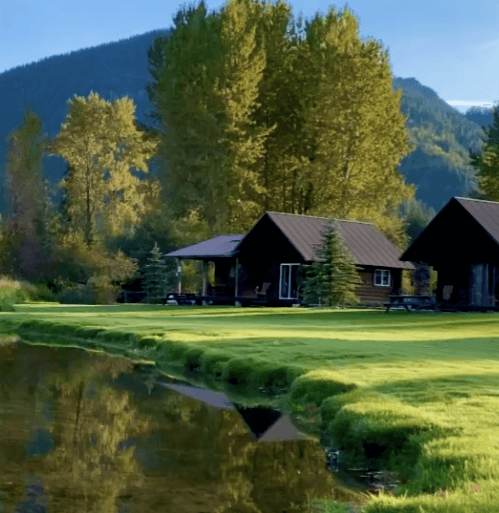 In the foreground, a pond with green, grassy banks reflects leaves of gold. Two cabins stand in the background with trees and mountain framing the landscape.