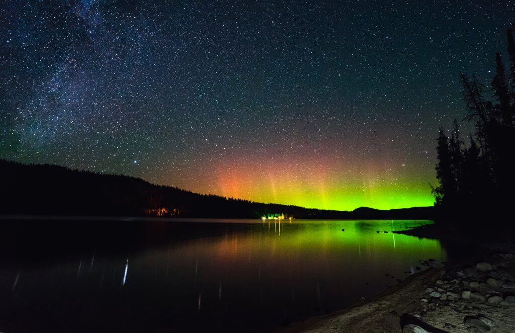 Green, orange and red northern lights over Redfish Lake at night with sparking stars of the Milky Way.