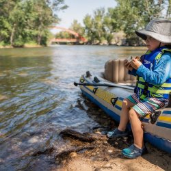 A small child sitting on a raft on the banks of the Boise River eating a sandwich.