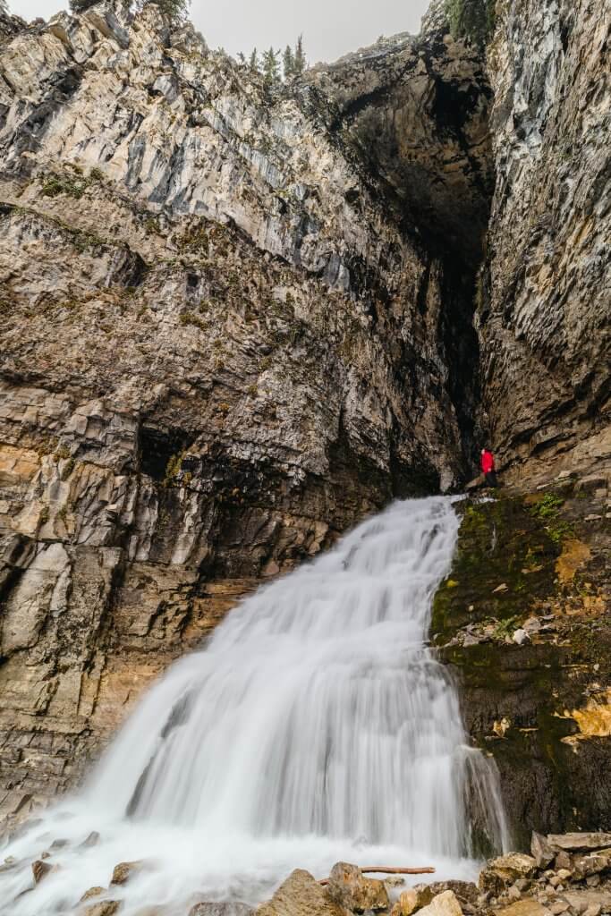 A man in a red jacket stands at the entrance to a cave where a waterfall is pouring out.