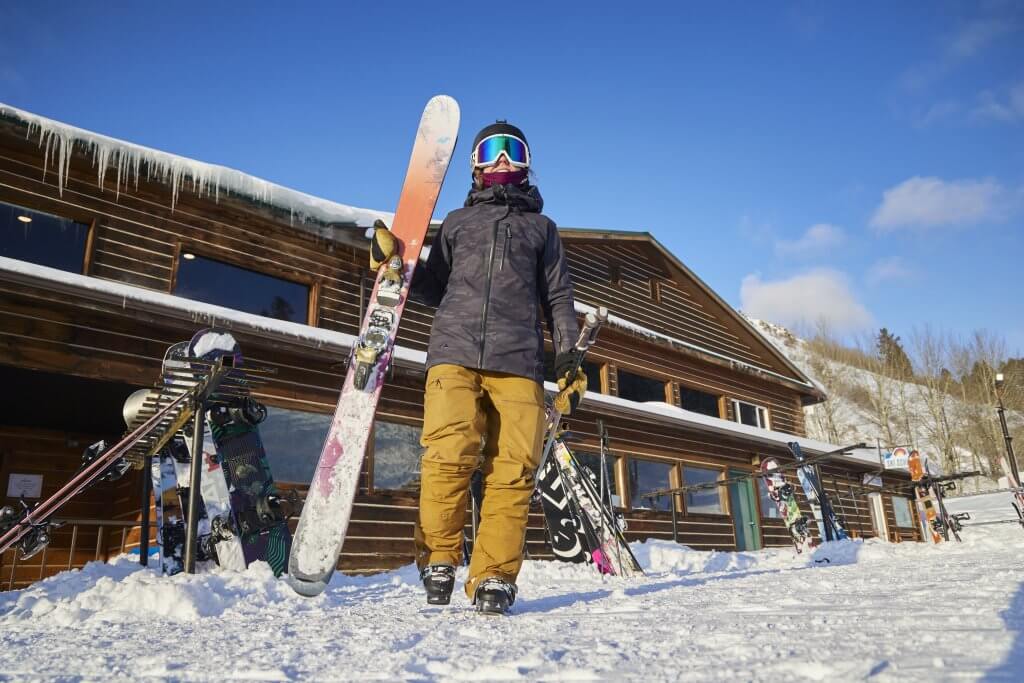 A person standing in front of a ski lodge at Kelly Canyon Resort holding a ski pole and a pair of skis.