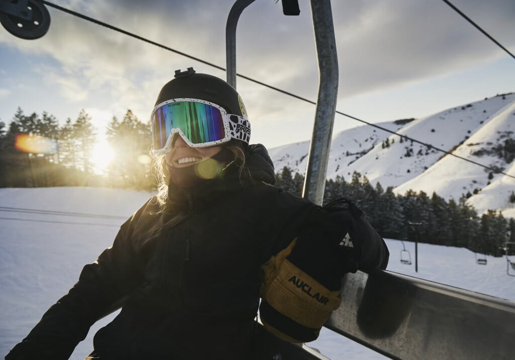 A woman wearing a helmet, goggles and a coat sitting on a ski lift, with snow-covered mountains and trees in the background, at Kelly Canyon Resort.