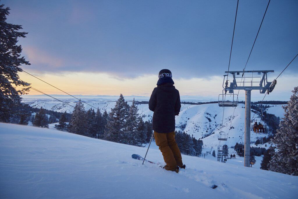 Backview of a person on skis at Kelly Canyon Resort, with a ski lift and snow-covered trees on either side of them and snow covered mountains in the distance.