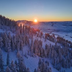 Sideview of a snow- and tree-covered mountain, with the sun rising over some mountains in the distance, at Pebble Creek Ski Area.