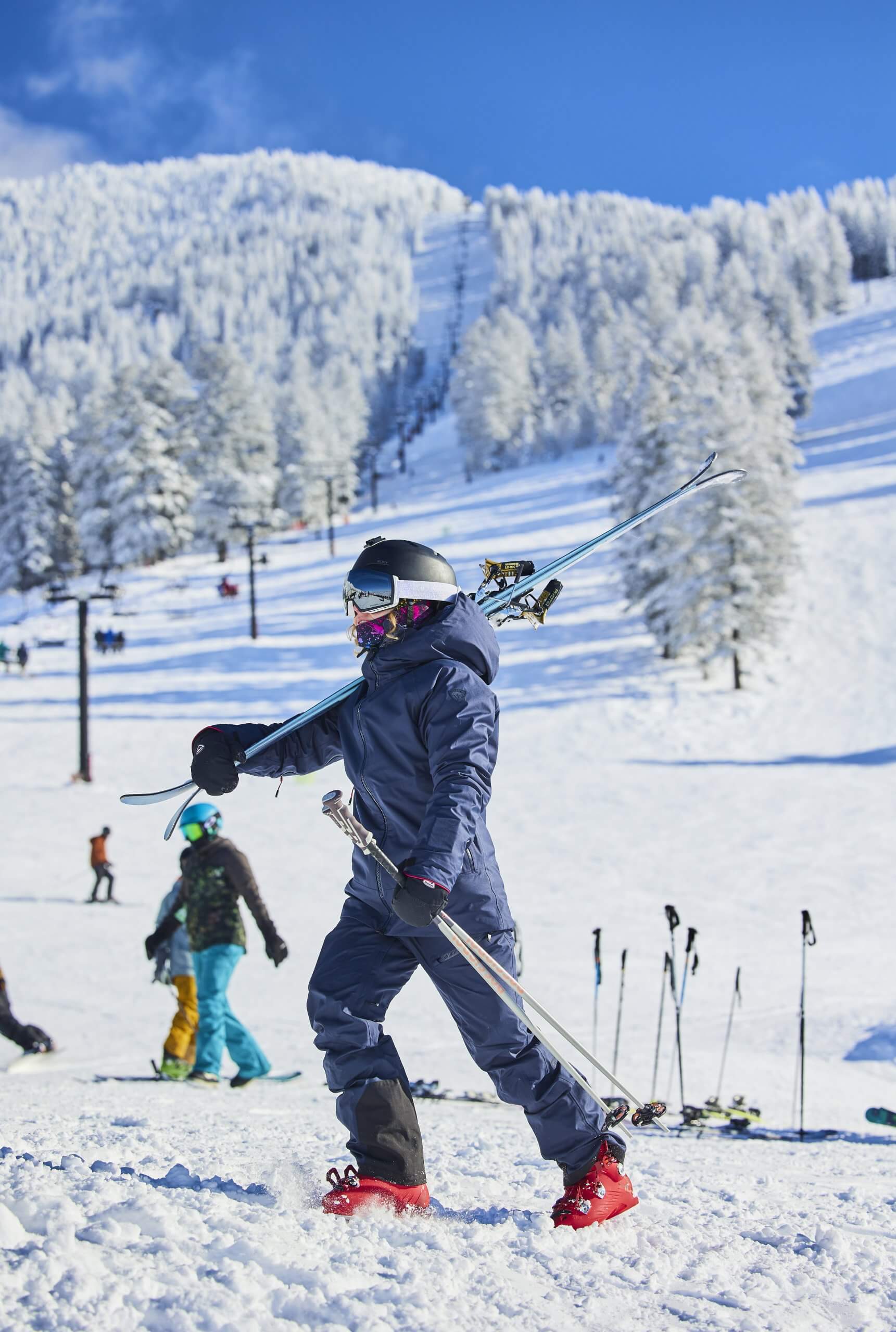 A person walking through the snow carrying ski equipment, and a forest of snow-covered trees in the background at Pebble Creek Ski Area.