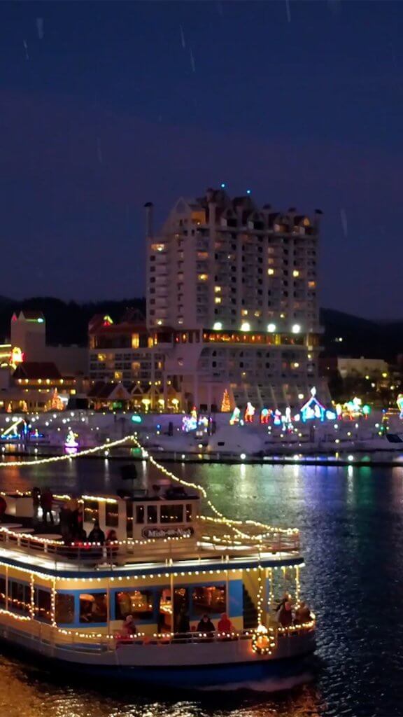 A cruise ship decorated with festive lights on Lake Couer d'Alene, with the city of Coeur d'Alene lit up with lights in the background. 