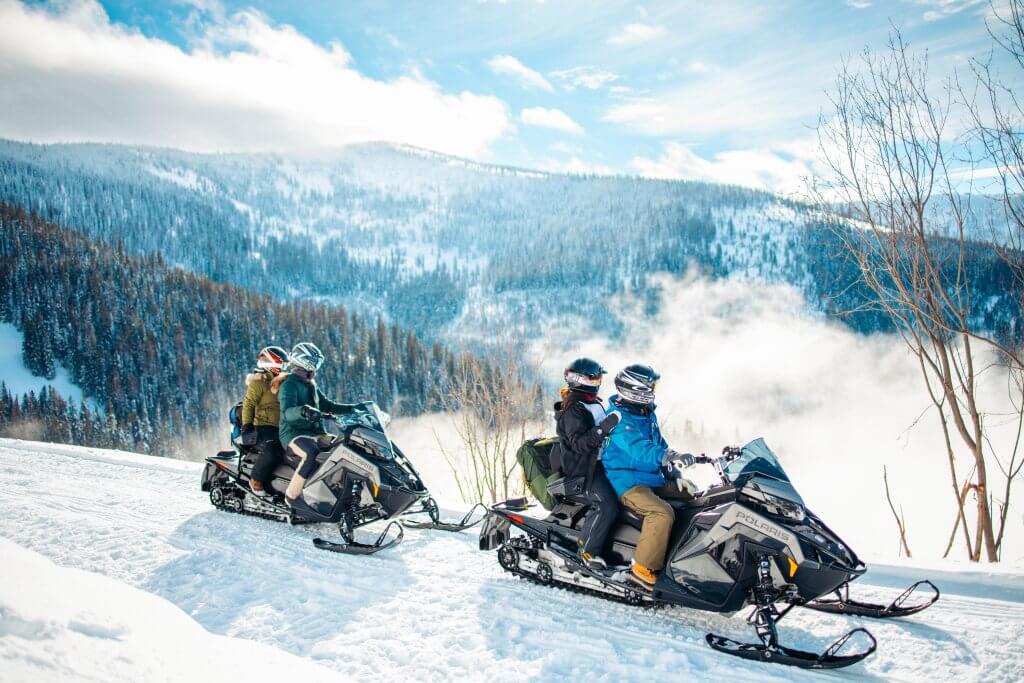 Two groups of people sitting on parked snowmobiles overlooking a scenic vista of a snow-covered forest in Sandpoint.