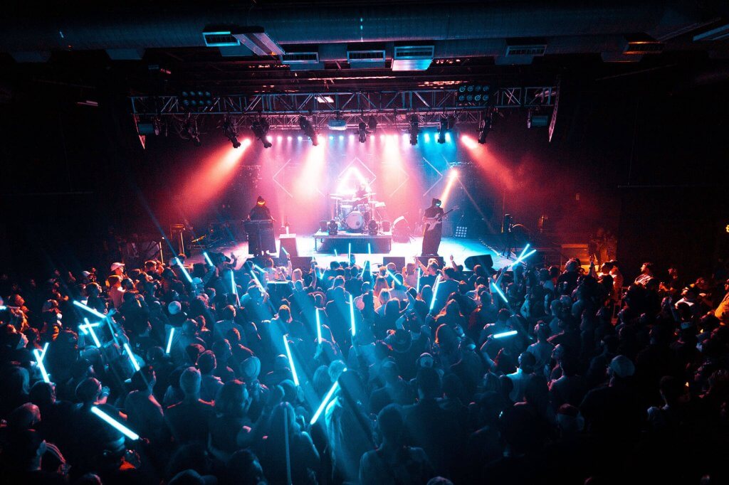 The band Magic Sword performing at Treefort Music Fest on a stage with colorful lights in front of a crowd holding long glow sticks.