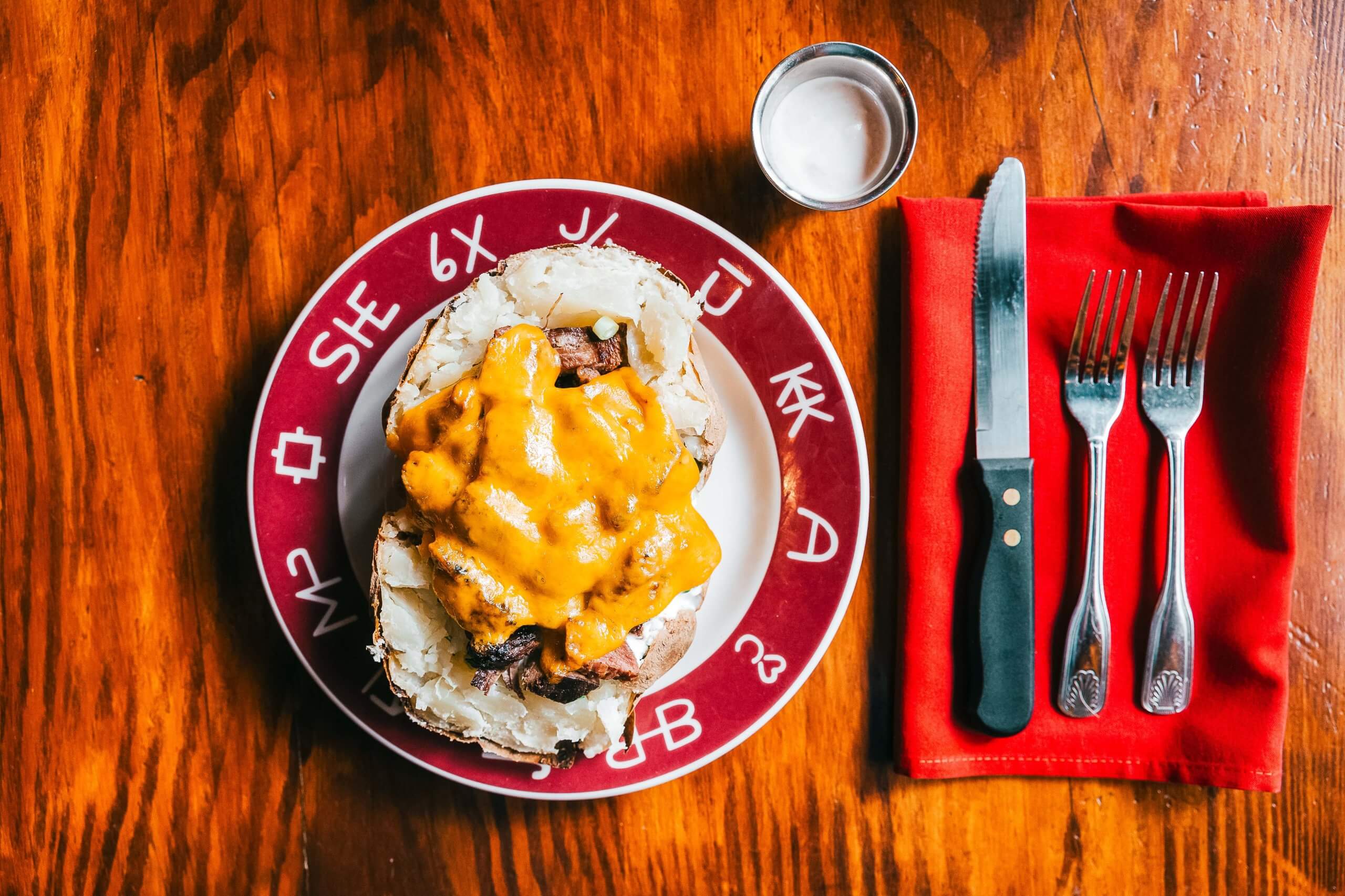 An overhead view of a Jim Spud, a large baked potato stuffed with meat and cheese, on a plate beside cup of sauce and cutlery on a table.