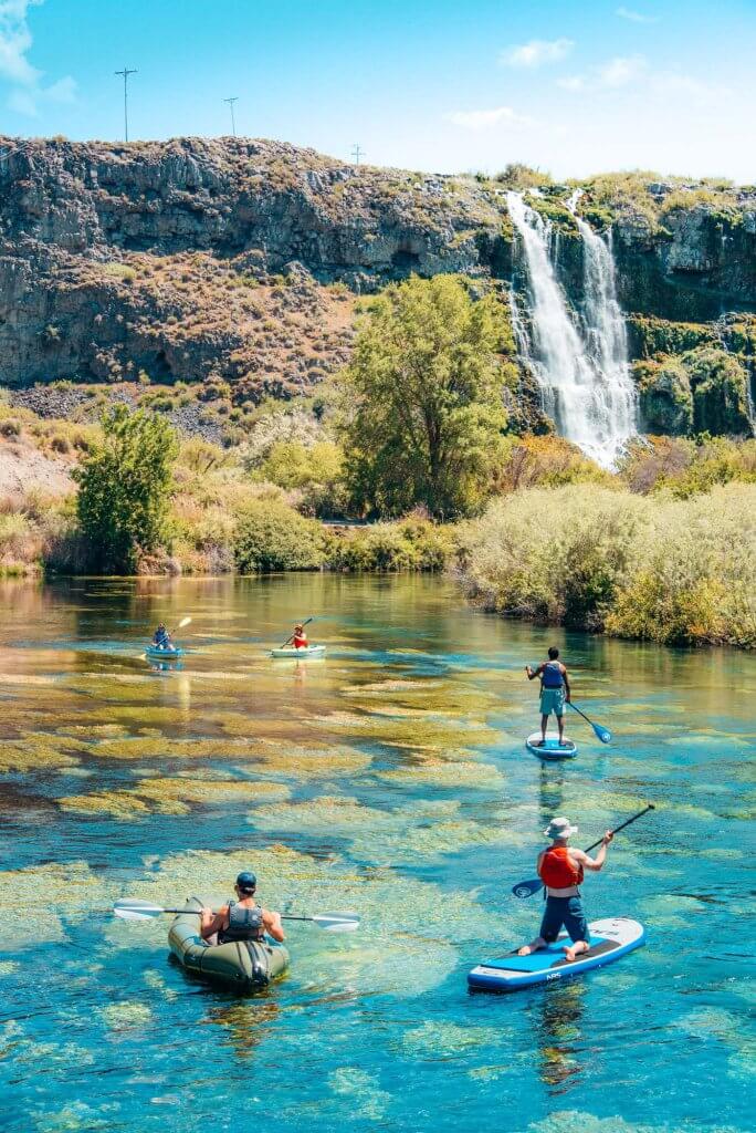 Five people float on kayaks and paddle boards on turquoise-blue water in front of a waterfall.