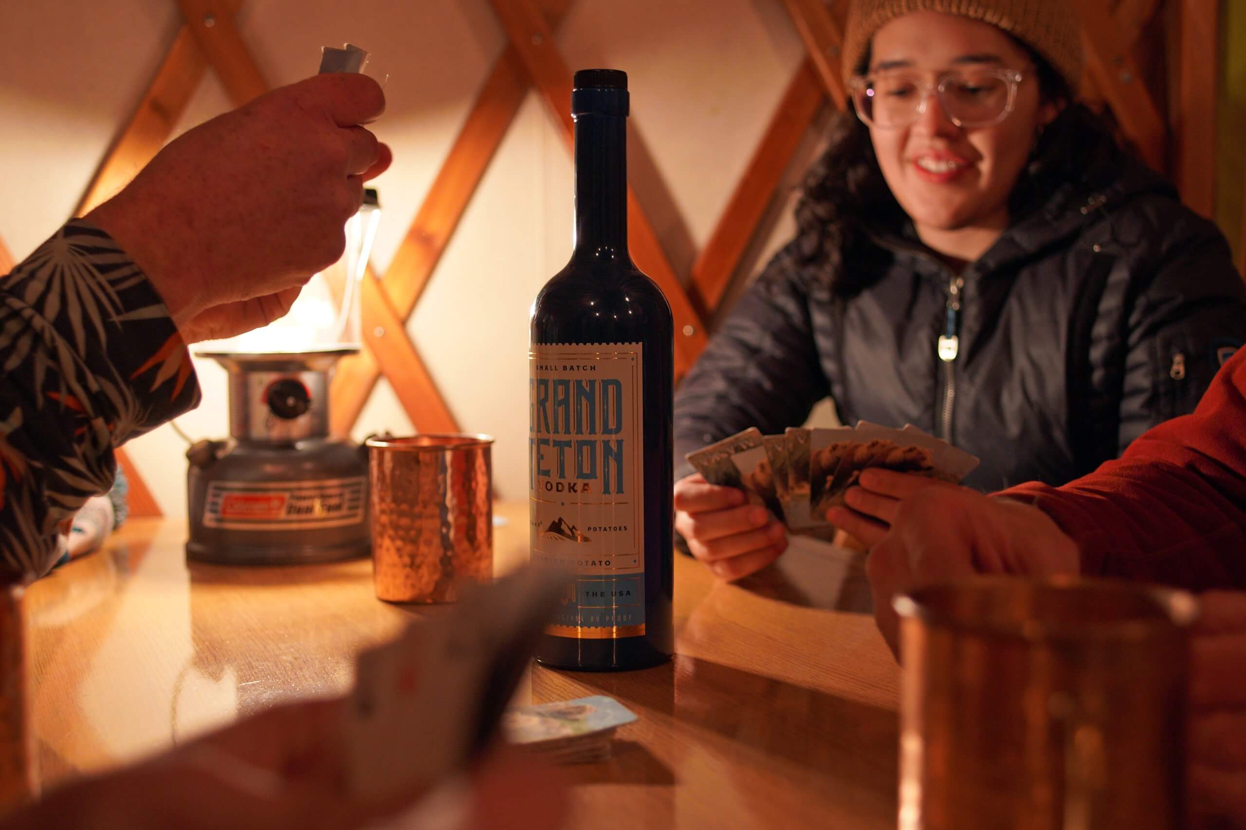 A bottle of Grand Teton Potato Vodka at the center of a table surrounded by a group of people playing cards.