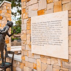 A statue of Anne Frank balancing on a chair looks out a window next to an interpretive sign.