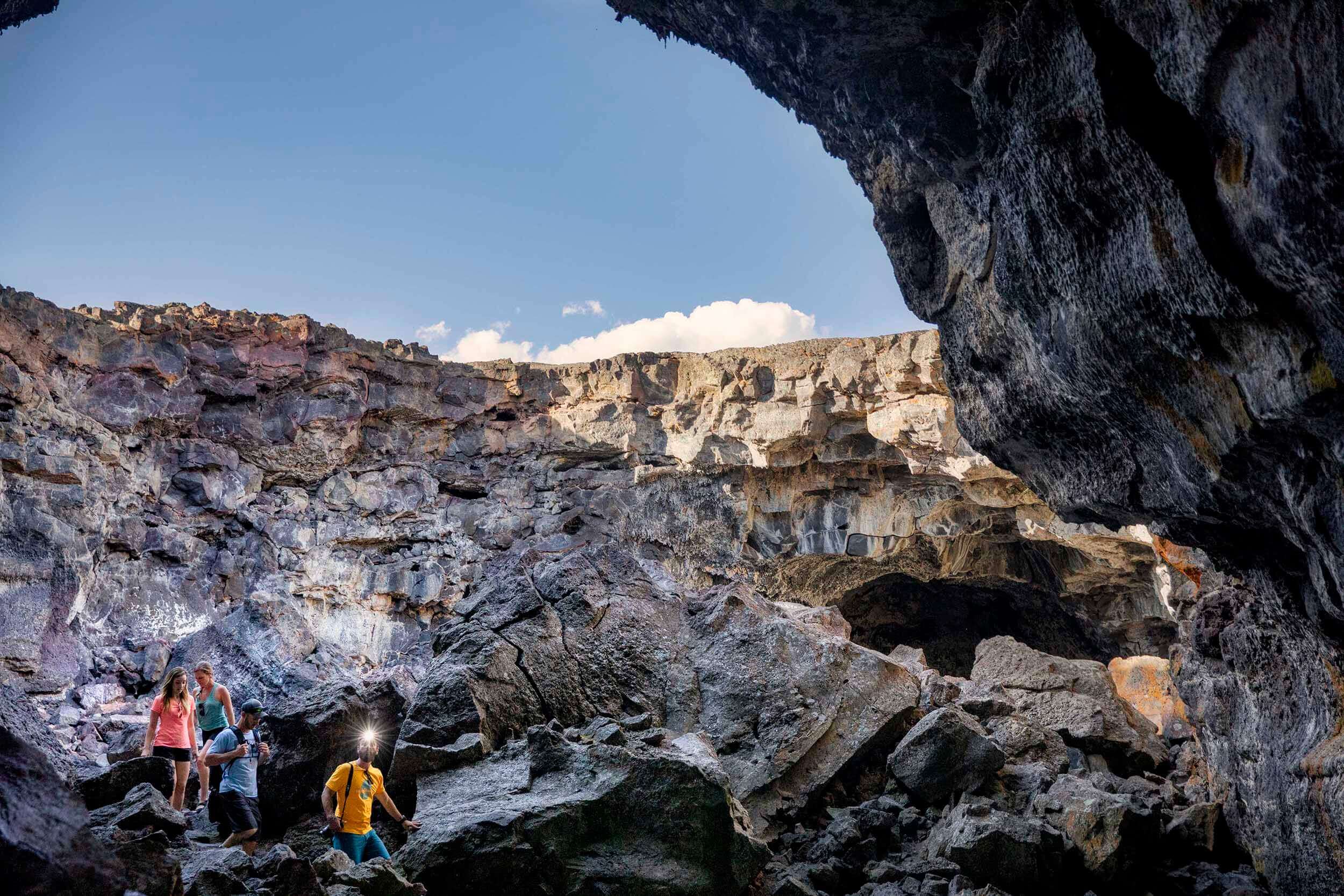 A group of four hike down an open lava tube toward a cave area.