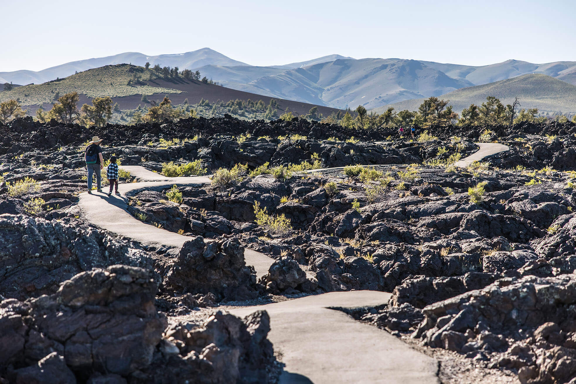 A man and child walk together on a paved trail through lava rock formations with mountains in the distance.