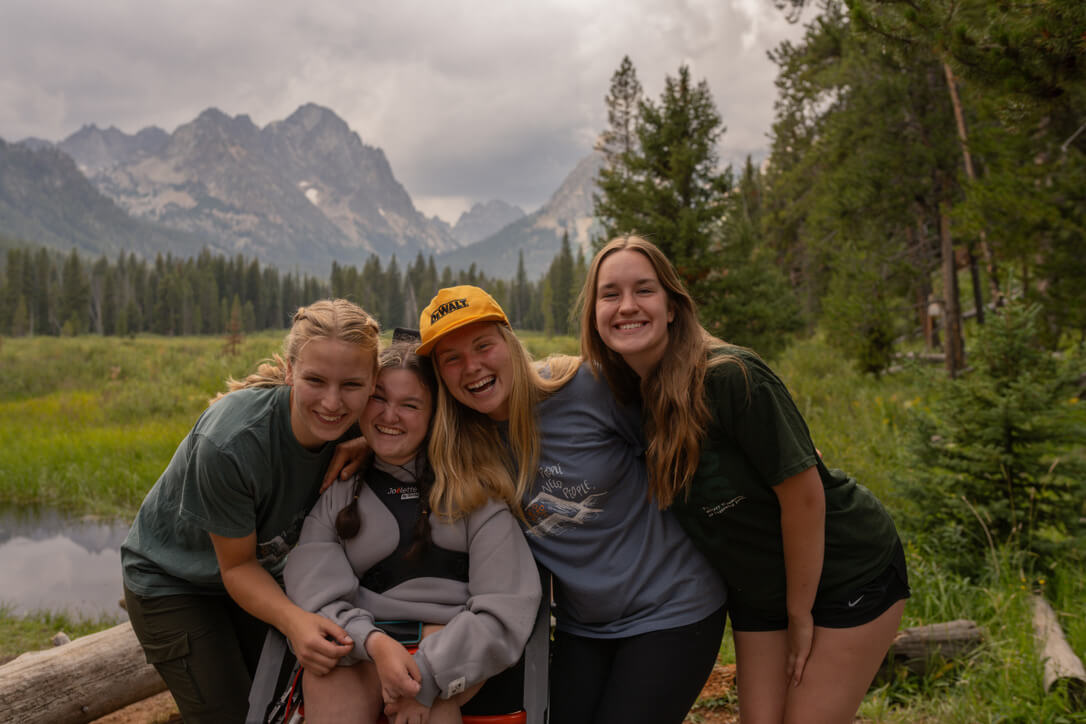 Four females are in a group, one of them is in an adaptive all-terrain wheelchair and they are smiling with the Sawtooth Mountains in the background.