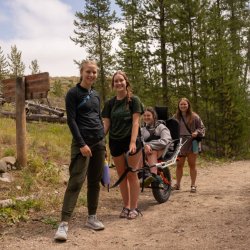 Four women are gathered near a trail sign, one of the women is in an adaptive all-terrain wheelchair.