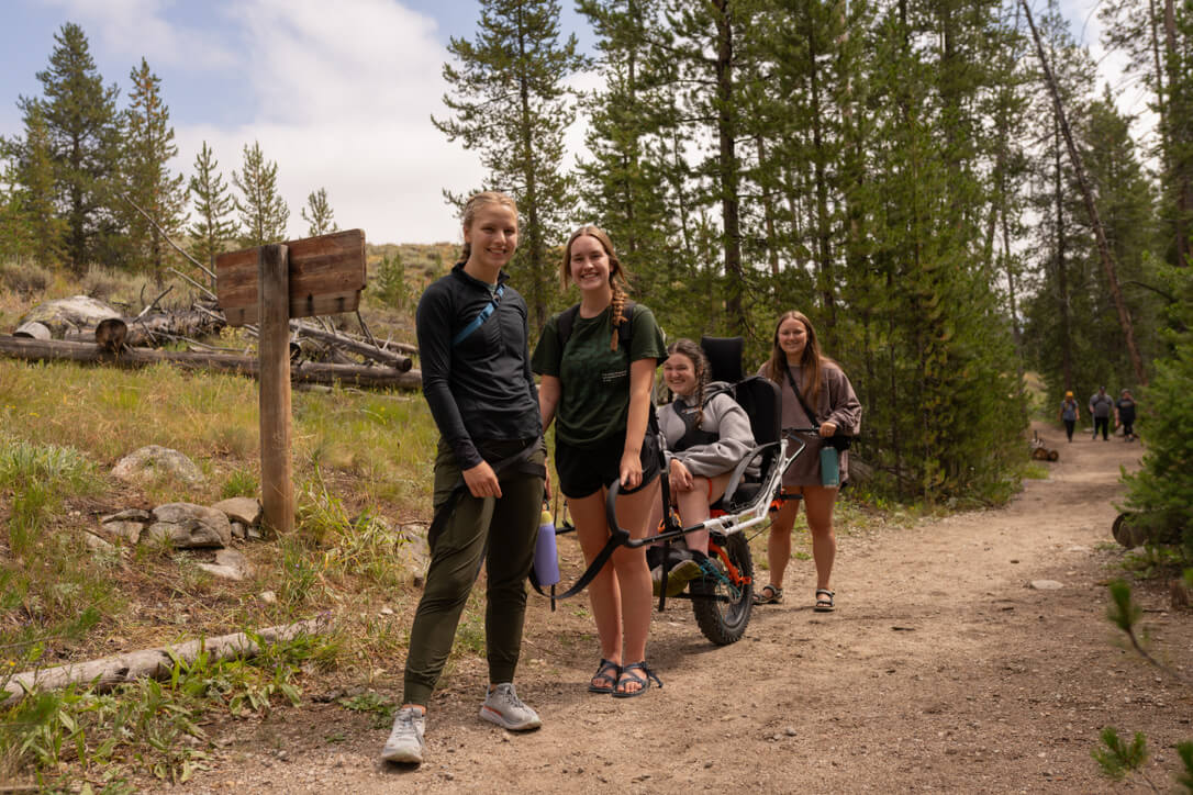 Four women are gathered near a trail sign, one of the women is in an adaptive all-terrain wheelchair.