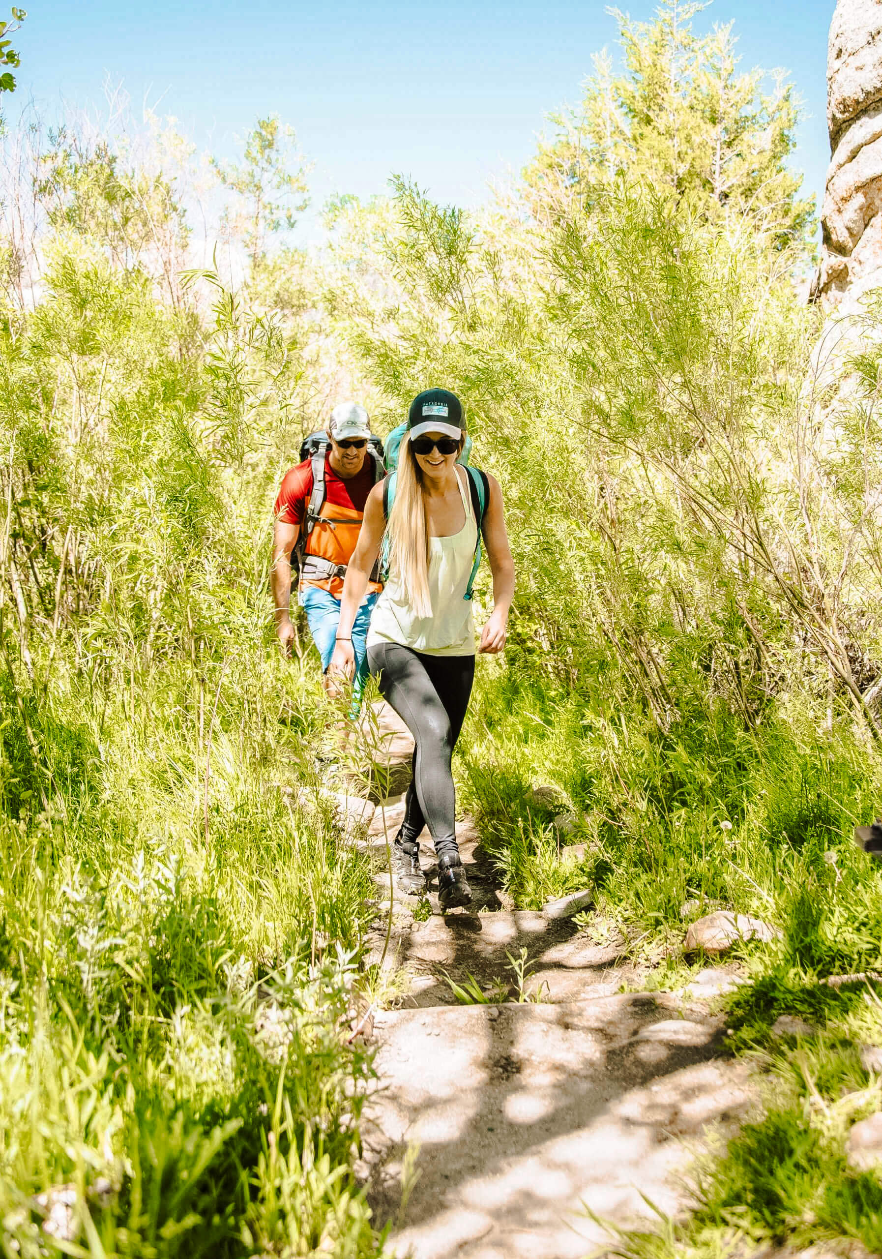 Two people hiking on a trail surrounded by tall shrubs and grass at City of Rocks National Reserve in the summer.